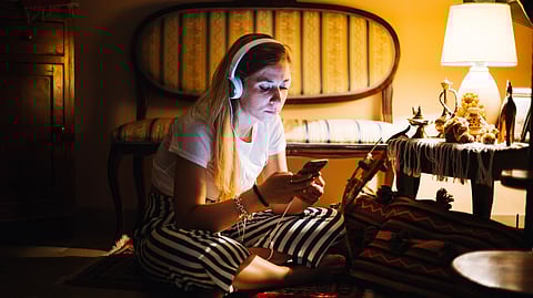A woman sits on the floor in a cozy, dimly lit room, wearing headphones and looking at her phone.