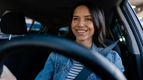 A woman in a denim jacket smiles while driving a car.