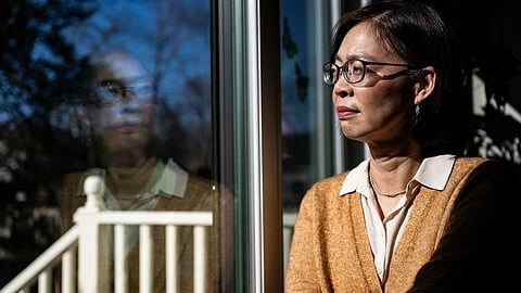 A woman with glasses and a brown cardigan gazes thoughtfully out a window. 