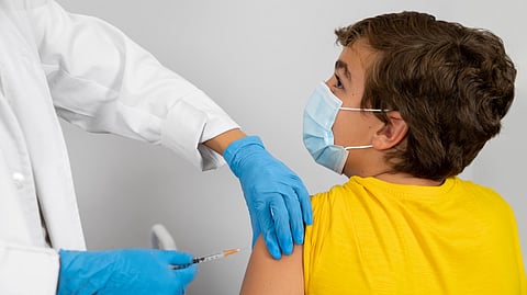 A child in a yellow shirt and face mask receives a vaccination in the arm.
