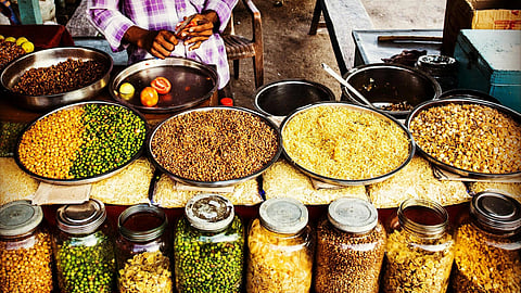 A street vendor arranges bowls filled with colorful grains and legumes, like chickpeas and lentils. Below, jars contain various dried snacks.
