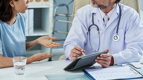 A doctor with a stethoscope and a clipboard is discussing with a patient in a blue shirt.