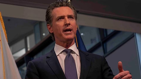 An image of Gavin Newsom wearing a suit, speaking at a podium with a confident expression. Behind him is a flag featuring a bear and a red star.