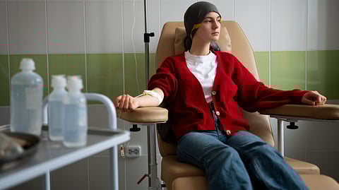 A woman in a red cardigan and headscarf sits in a hospital chair receiving treatment, with IV drip beside her.