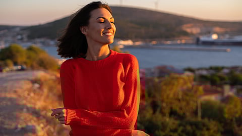 Woman in a red sweater smiling with eyes closed, stands in a sunlit landscape.