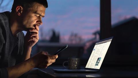 Man focused on a smartphone and laptop in a dimly lit room.
