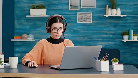 Woman in orange sweater and glasses focuses on laptop, wearing headphones.
