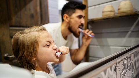 A child and an adult brush their teeth energetically in a bathroom, reflecting in a mirror.