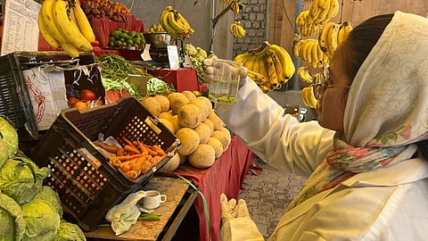 Woman in lab coat and headscarf checks green liquid at a market with fruits and vegetables around.