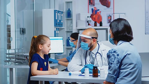 A doctor in a face shield and mask, seated at a desk with a stethoscope, attentively talks to a young girl in a clinic.