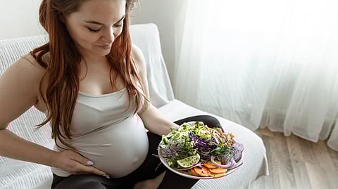 A pregnant woman sits on a couch, holding a plate of fresh salad with greens, sliced vegetables, and avocado.