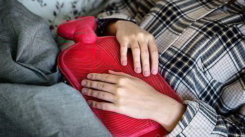 A person lies in bed wearing plaid pajamas, holding a red hot water bottle on their stomach. 