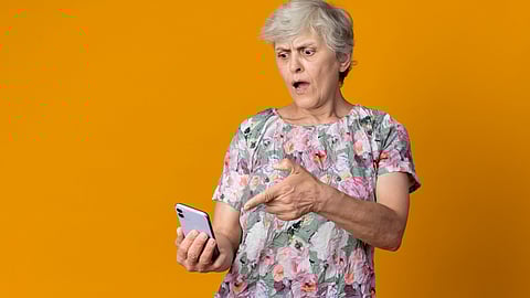 Elderly woman with short grey hair looks surprised, pointing at a smartphone. She wears a floral blouse against an orange background.