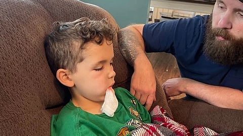 A young boy rests on a couch with gauze on his chin while a man sits beside him for support. 