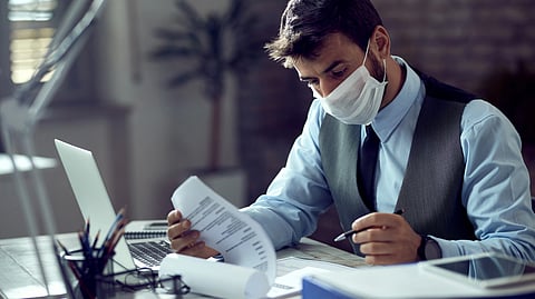 A man wearing a face mask and formal attire works intently at a desk, reviewing documents beside a laptop. 