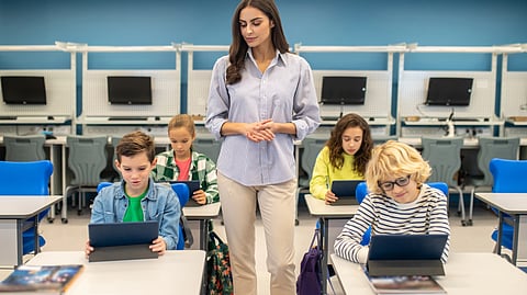A teacher observes as four students use tablets in a modern classroom with computers on desks.