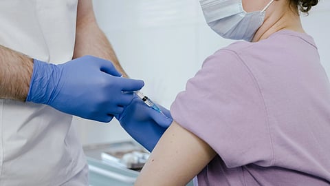 Healthcare worker in blue gloves administers a vaccine to a person wearing a lavender shirt and mask.