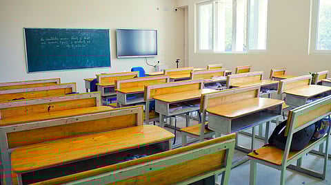 Empty classroom with wooden desks and chairs, a green chalkboard with writing.