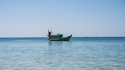 A small, colorful fishing boat with red flags floats on a calm, clear blue sea under a cloudless sky.
