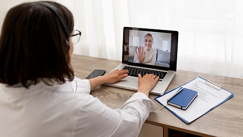 A woman in a white coat engages in a video call on a laptop, smiling person waves back. 