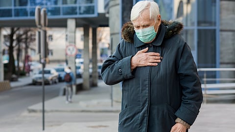 Elderly person with white hair wearing a mask and dark coat, touching chest.