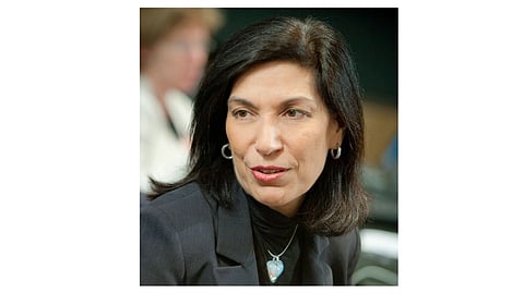 A woman with shoulder-length dark hair is speaking, wearing a black blazer, silver hoop earrings.