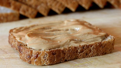 Slice of multigrain bread topped with creamy peanut butter on a light wood surface.