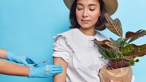 A person in a white blouse and straw hat holds a potted plant while receiving a vaccine.