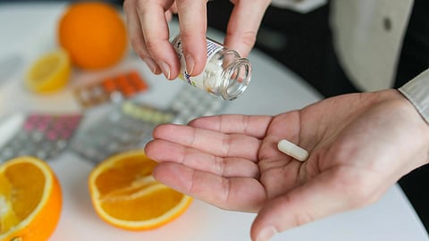 A person pours a white capsule from a bottle into their hand. An open orange, various pill packs.