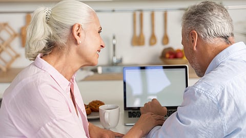Elderly couple sitting at a kitchen table, smiling and focused on a laptop screen. 