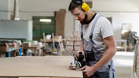 A worker cuts a board with a circular saw in a workshop while wearing safety gear.