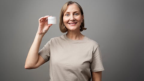 A smiling woman in a beige shirt holds up a small white cream jar against a gray background.