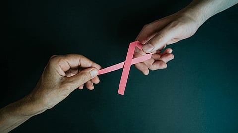 Two hands hold a pink ribbon against a dark background.