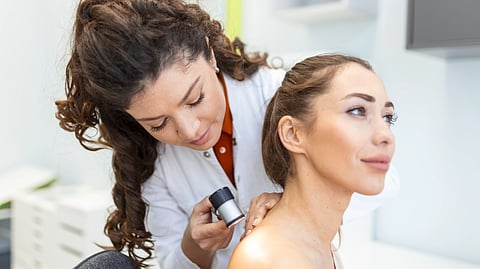 A dermatologist in a white coat examines a woman’s shoulder with a dermatoscope.