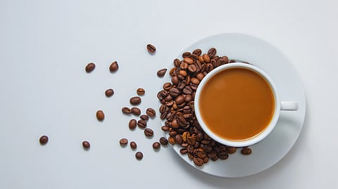 A white cup filled with coffee sits on a saucer. Coffee beans scatter to the left.