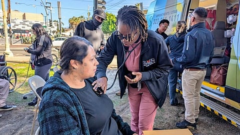 A healthcare worker checks a seated woman's vitals outdoors near a mobile clinic van. 