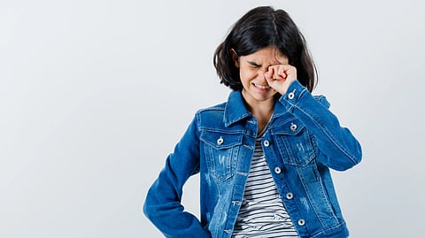 A woman in a denim jacket and striped shirt rubs her eye, smiling slightly. 