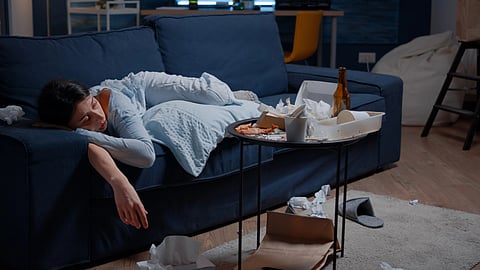 A woman in pajamas sleeps on a blue couch, surrounded by messy food containers and tissues on a cluttered table.