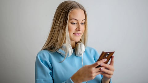 Young woman in a blue sweater with headphones around her neck, looking at her phone with a slight smile. 
