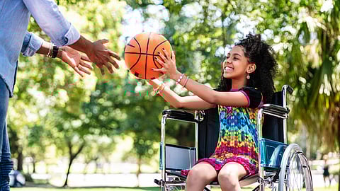 A smiling young girl in a wheelchair catches a basketball passed by an adult.