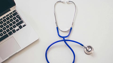 A stethoscope with a blue tube is coiled on a white surface next to an open silver laptop. 
