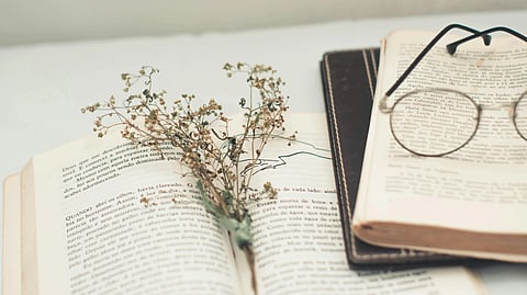 Open books with text in a soft focus, a sprig of dried flowers placed inside one, and round glasses resting on another.
