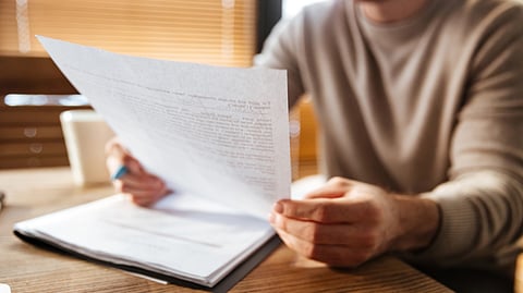 A person in a beige sweater reviews documents at a wooden table.