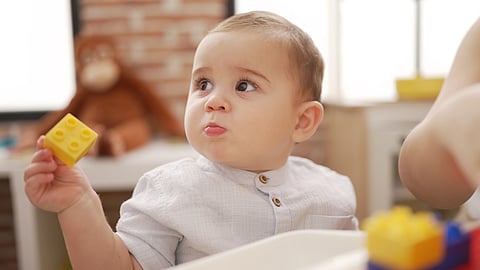 A baby wearing a light shirt holds a yellow building block.