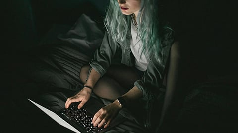 A person with long, teal hair types on a laptop in a dimly lit room, sitting cross-legged on a bed.