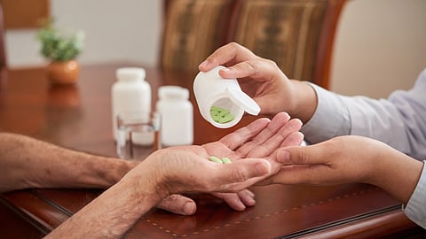 Two hands transfer green pills from a bottle into an elderly person's hand at a wooden table.