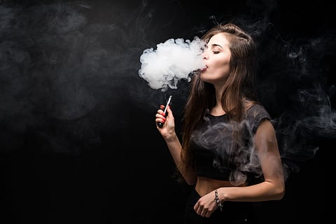 Young woman in black smokes an electronic cigarette on dark wall