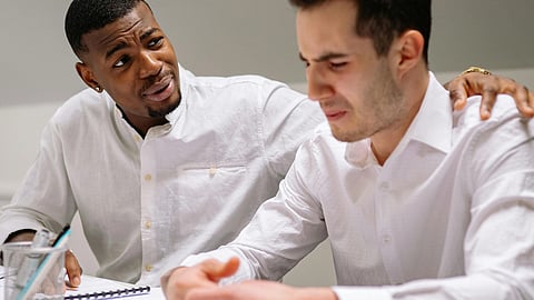 A man comforting a confused coworker at a desk, both wearing white shirts, with papers and pens around.