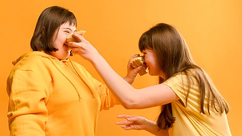 Two women in yellow outfits laugh as they playfully press slices of cake on each other's faces against an orange background.