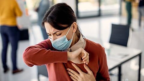 A woman wearing a face mask coughs into her elbow in a bright office, conveying an awareness of health precautions.